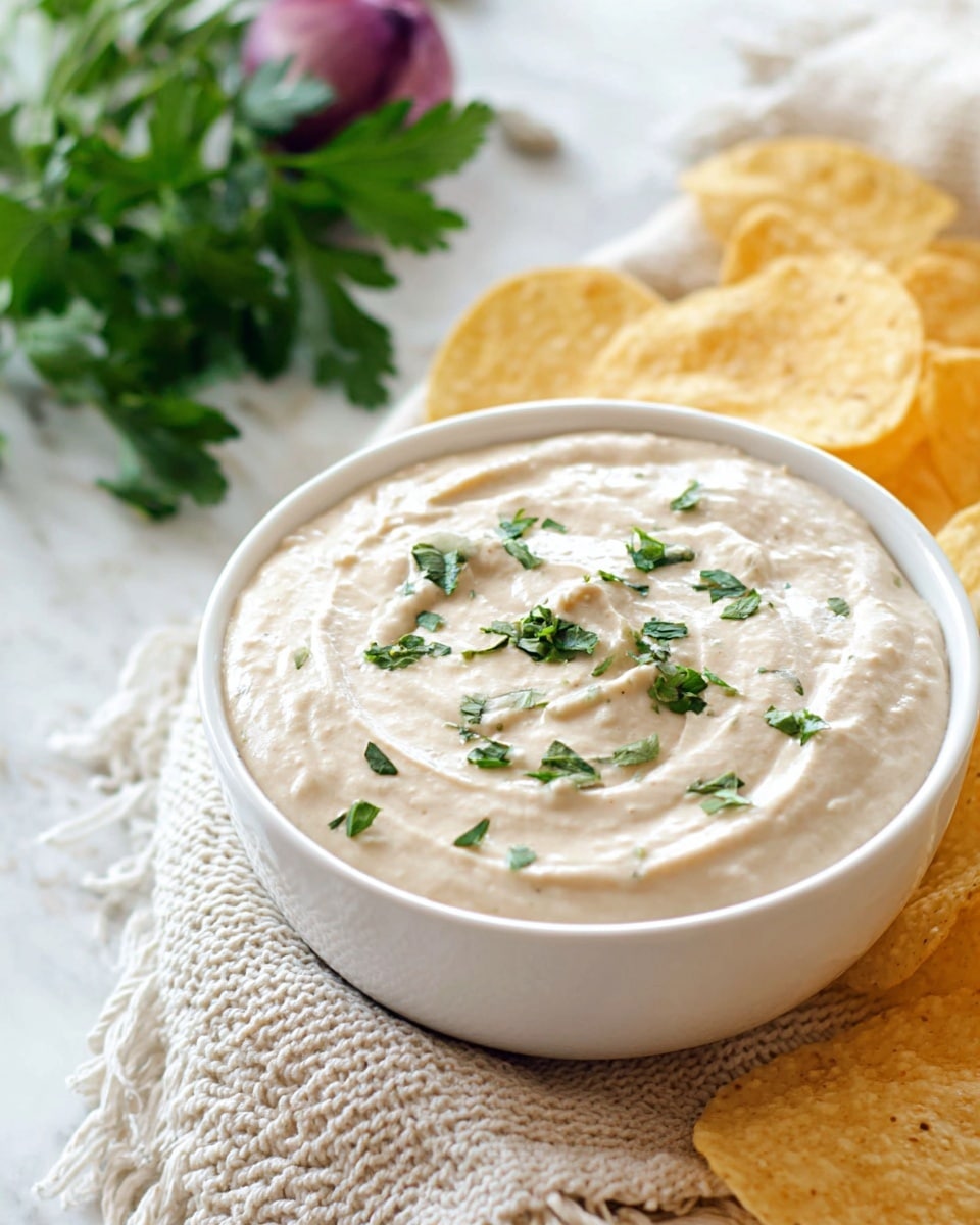 A white bowl filled with smooth, creamy light beige dip decorated with small pieces of green herbs on top, placed on a white marbled surface with several round, light yellow tortilla chips partially visible behind the bowl, and a bunch of fresh green parsley and a purple onion peeking from the bottom edge, with a knitted light beige cloth nearby. Photo taken with an iphone --ar 4:5 --v 7