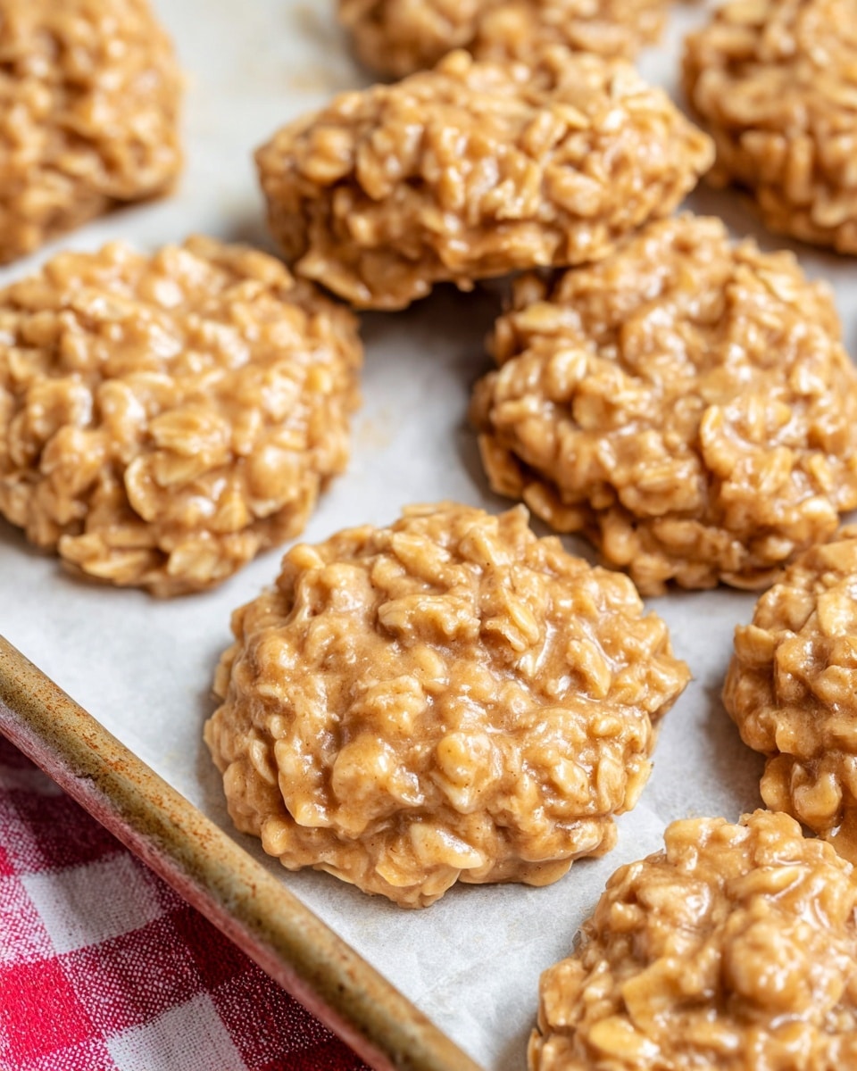 The image shows several round oatmeal cookies in a close-up view. Each cookie has a rough texture with visible oats embedded in a light brown, slightly shiny dough. The cookies are arranged on white parchment paper that lines a metal baking tray. The tray rests on a white marbled surface with a red and white checkered cloth visible on one corner. The cookies overlap slightly, showing uneven edges and a homemade look. Photo taken with an iphone --ar 4:5 --v 7