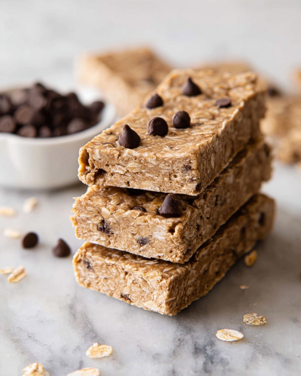 A stack of four rectangular oat and chocolate chip bars is placed on a white marbled surface. The bars have a light brown color with visible oat flakes mixed throughout. Small dark chocolate chips are embedded inside the bars, while a few are scattered on top and around the stack. In the background, a small white bowl filled with chocolate chips and a few more bars are slightly blurred. The texture of the bars looks soft and dense, and the light creates a natural shine on their surface. photo taken with an iphone --ar 4:5 --v 7