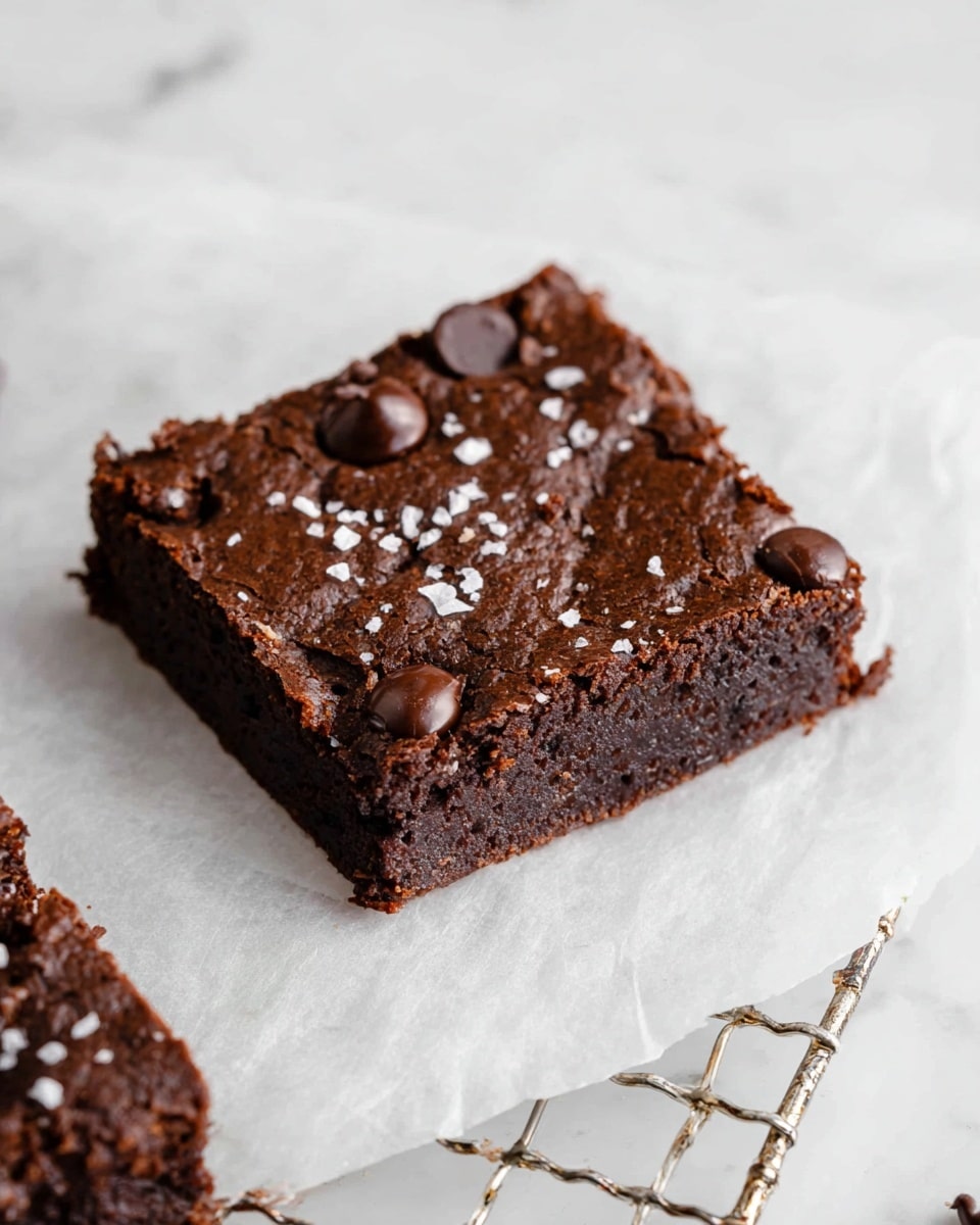 A thick, dark brown chocolate brownie square sits on white parchment paper placed on a wire cooling rack with a metallic shine. The brownie has a slightly cracked surface texture with visible glossy chocolate chips scattered on top, and a light sprinkling of coarse white sea salt flakes. The edges of the brownie look firm while the center is dense and moist. The background is a white marbled texture with faint patterns. photo taken with an iphone --ar 4:5 --v 7
