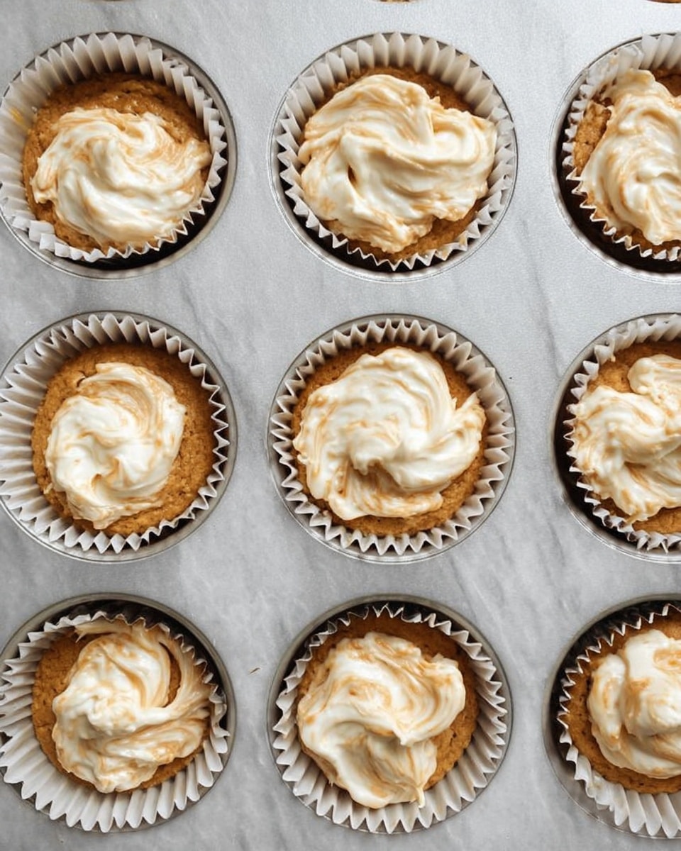 The image shows a metal muffin tray with nine white paper cupcake liners filled with light brown cupcake batter. Each cupcake has a swirl of creamy white frosting or cream cheese topping in the center, slightly mixed into the batter creating a marbled effect. The tray sits on a white marbled surface, and the texture of the batter looks smooth but thick, while the topping is soft and creamy. The cupcakes are evenly spaced in the tray with a close-up overhead view. Photo taken with an iphone --ar 4:5 --v 7