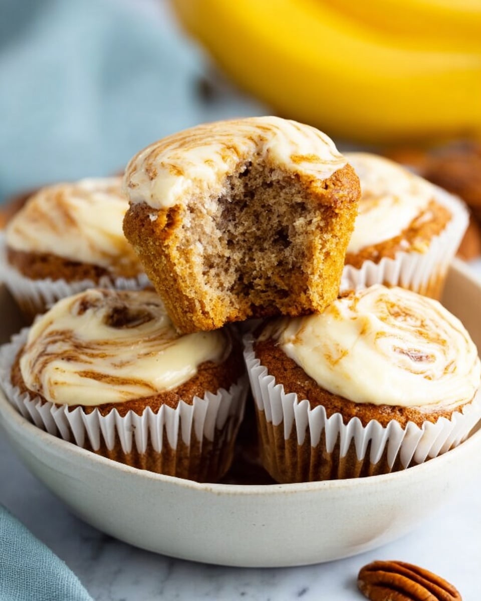 A close-up bowl filled with five muffins, one muffin held above the others showing a bite taken out of it, revealing a soft, light brown interior with darker specks inside. Each muffin has a top layer of creamy white frosting swirled with light brown streaks. The muffins are wrapped in white paper liners. The bowl is white and sits on a white marbled surface, with a few pecan nuts placed next to the bowl. In the background, a blurred banana adds a splash of yellow color. photo taken with an iphone --ar 4:5 --v 7