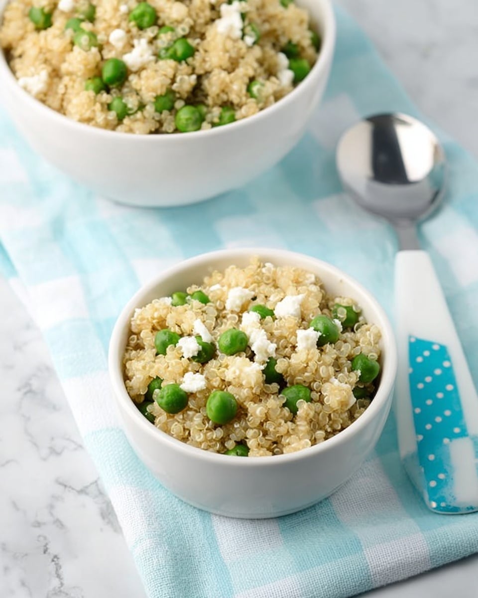 The image shows two white bowls filled with a mix of light beige quinoa and bright green peas, with small white crumbles of cheese scattered on top. The bowl in the front is smaller and centered, with the quinoa and peas visibly mixed with crumbled cheese pieces evenly spread. Behind it, a larger bowl also contains the same mixture. The bowls sit on a light blue and white checkered cloth, placed on a white marbled surface. To the right, there is a spoon with a white tip and a blue handle with white polka dots. Photo taken with an iphone --ar 4:5 --v 7