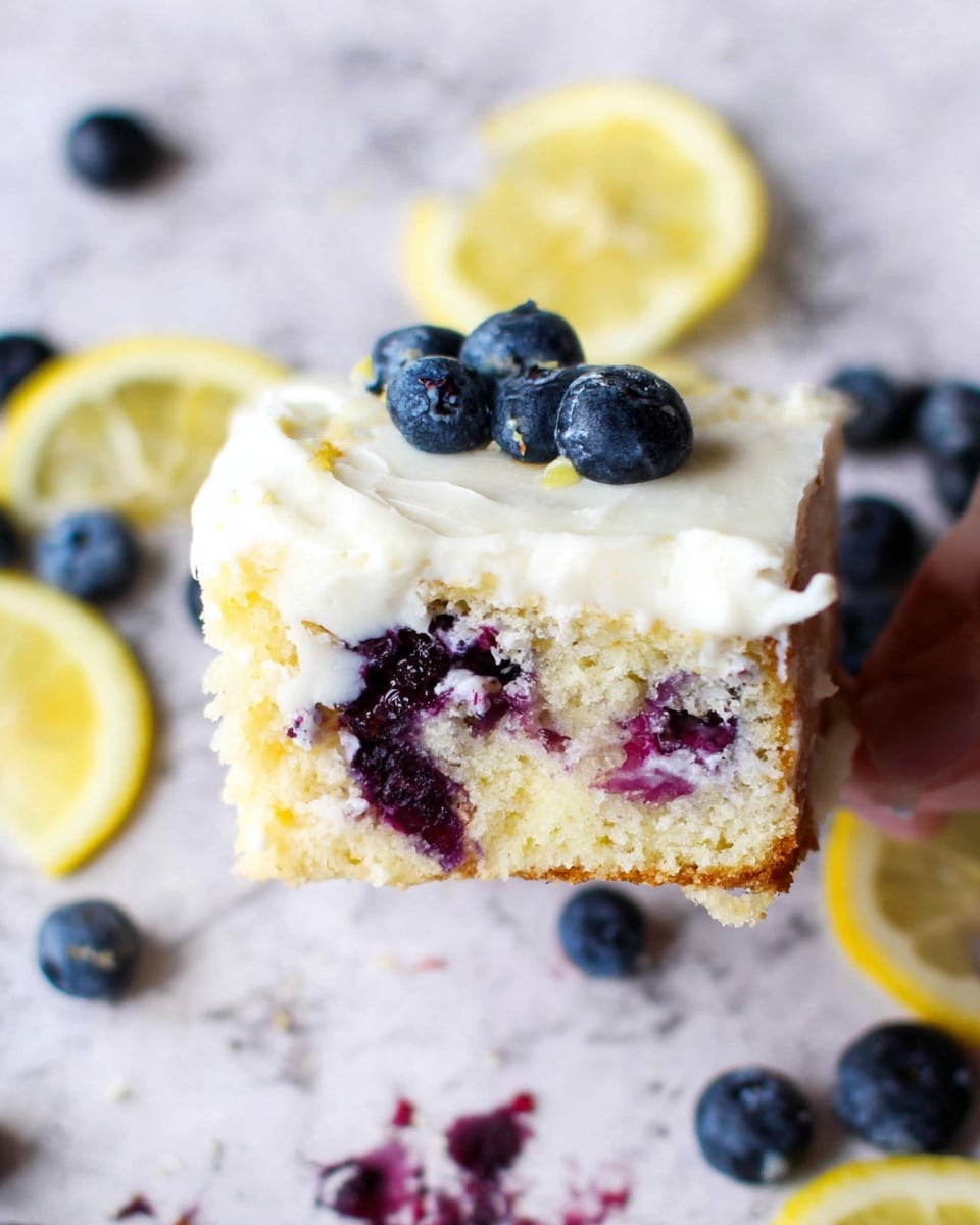 The image shows a piece of blueberry lemon cake held by a woman's hand. The cake has two visible layers: the bottom layer is a light yellow, soft sponge dotted with whole and mashed blueberries that create deep purple spots. The top layer is a smooth, thick white frosting with some blueberries pressed into it. Around the cake slice lie loose blueberries and thin lemon slices scattered on a white marbled surface, adding bright blue and yellow pops of color to the scene. The texture of the cake looks moist and fluffy, while the frosting is creamy and smooth. Photo taken with an iphone --ar 4:5 --v 7