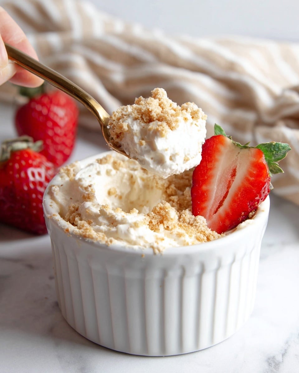 A white ribbed ramekin dish filled with a creamy white mixture that has a soft texture and crumbly light brown bits sprinkled on top as one layer. Behind the ramekin are two red strawberries, one whole with green leaves and one sliced in half showing the bright red inside. A gold spoon held by a woman's hand lifts a scoop of the creamy mixture mixed with the light brown crumbs. The background has a white marbled texture with a beige striped cloth partially visible. Photo taken with an iphone --ar 4:5 --v 7