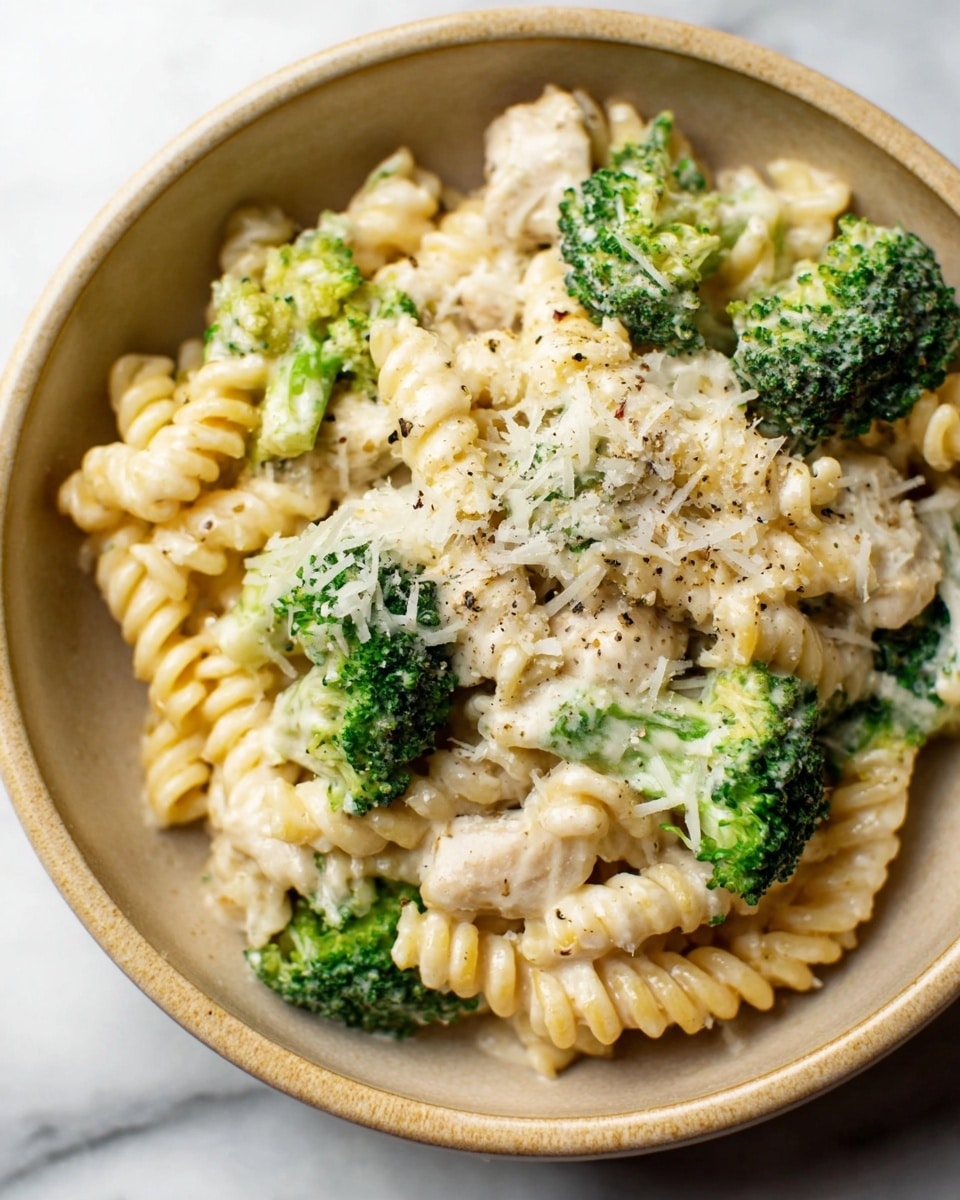 A close-up view of a bowl filled with creamy pasta made of spiral rotini noodles coated in a white cheese sauce. Scattered throughout the pasta are bright green broccoli florets that add a fresh pop of color. Small, tender pieces of white meat are mixed in, covered by the sauce. The dish is topped with finely grated cheese and small sprinkles of herbs and black pepper, all sitting in a beige ceramic bowl placed on a white marbled surface photo taken with an iphone --ar 4:5 --v 7