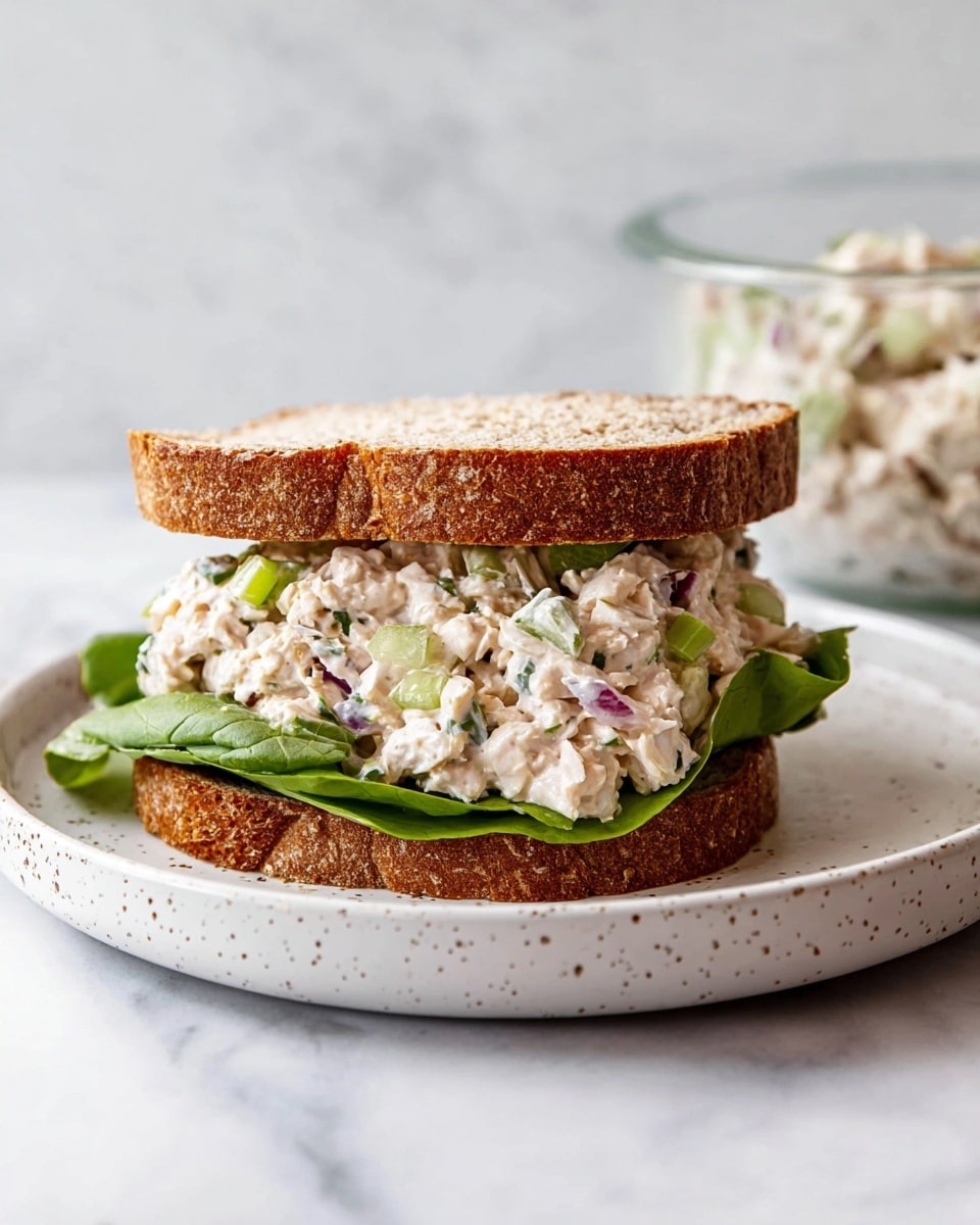 A sandwich with two slices of brown bread forms the top and bottom layers, with fresh green lettuce leaves lying directly on the bottom bread slice. On top of the lettuce, there is a thick layer of chunky white chicken salad mixed with small pieces of green celery and purple onion, creating a textured, colorful filling. The sandwich is placed on a white plate with a subtle speckled pattern, all set against a white marbled surface. In the background, there is a clear glass bowl partially visible with more of the chicken salad inside. Photo taken with an iphone --ar 4:5 --v 7