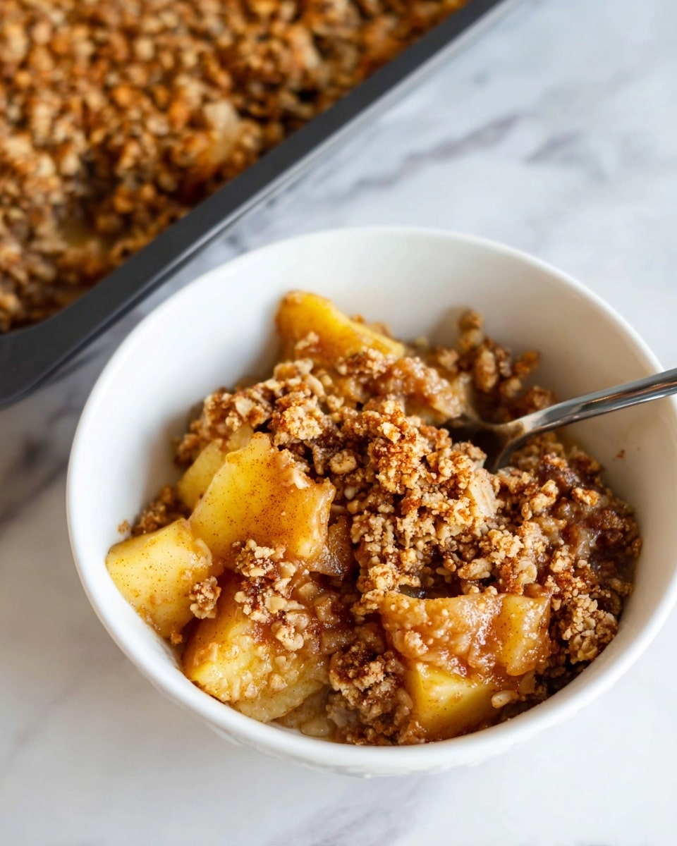 A white bowl holds a serving of apple crisp with two main layers visible; the bottom layer consists of soft, warm apple pieces in a light golden brown color, some showing a bit of cinnamon, while the top layer is a crumbly, textured topping that is golden brown with small clusters and a slightly crunchy appearance. A spoon is partially submerged in the bowl, and the bowl sits on a white marbled surface. In the background, a dark baking tray with more of the apple crisp is partially visible. photo taken with an iphone --ar 4:5 --v 7