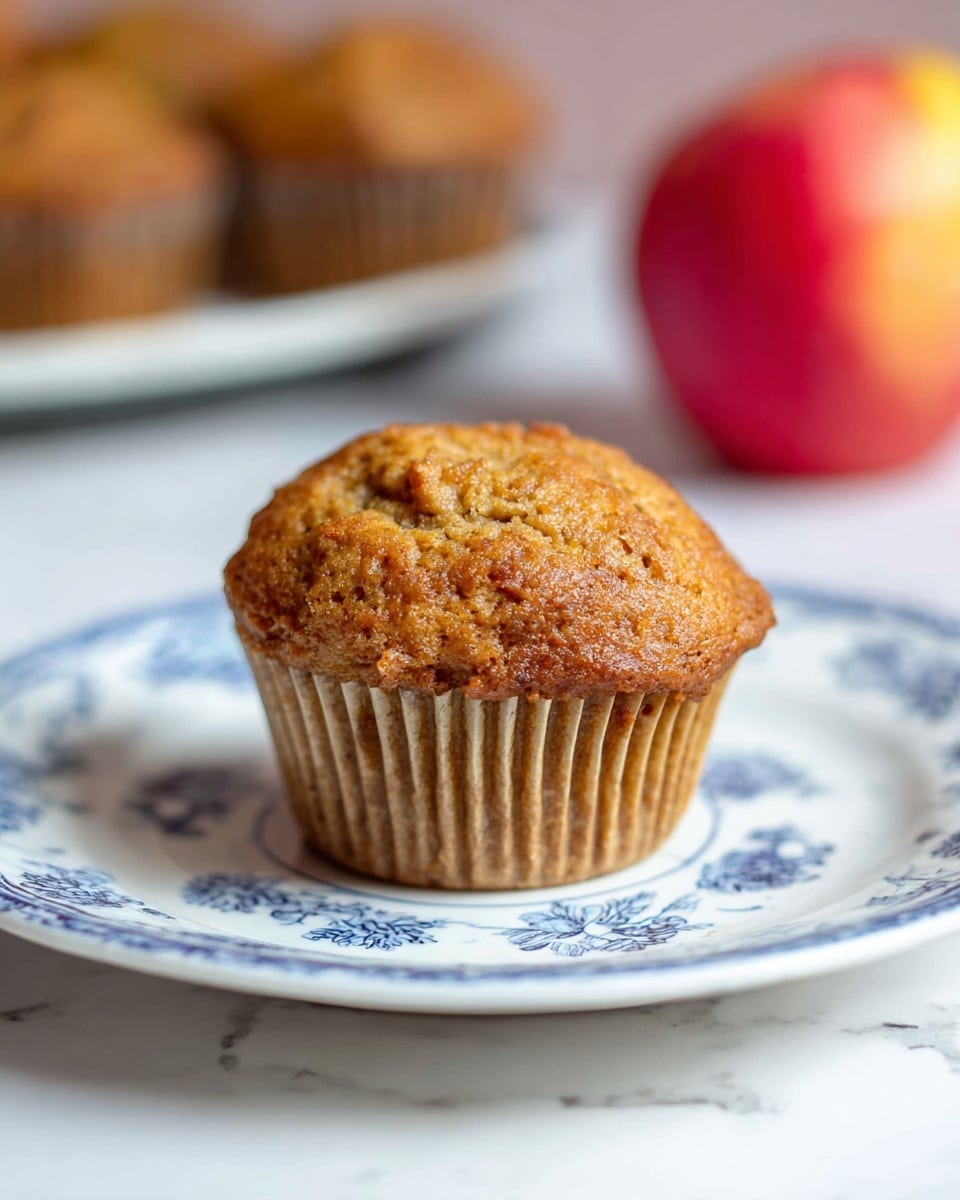 A single golden brown muffin with a slightly rough, textured top sits on a white plate decorated with intricate blue floral patterns. The muffin's paper liner is light brown and slightly wrinkled, hugging the base closely. The plate rests on a white marbled surface, and in the blurred background, there is a soft glimpse of a red and yellow apple on the right and a tray with more muffins on the left. Photo taken with an iphone --ar 4:5 --v 7