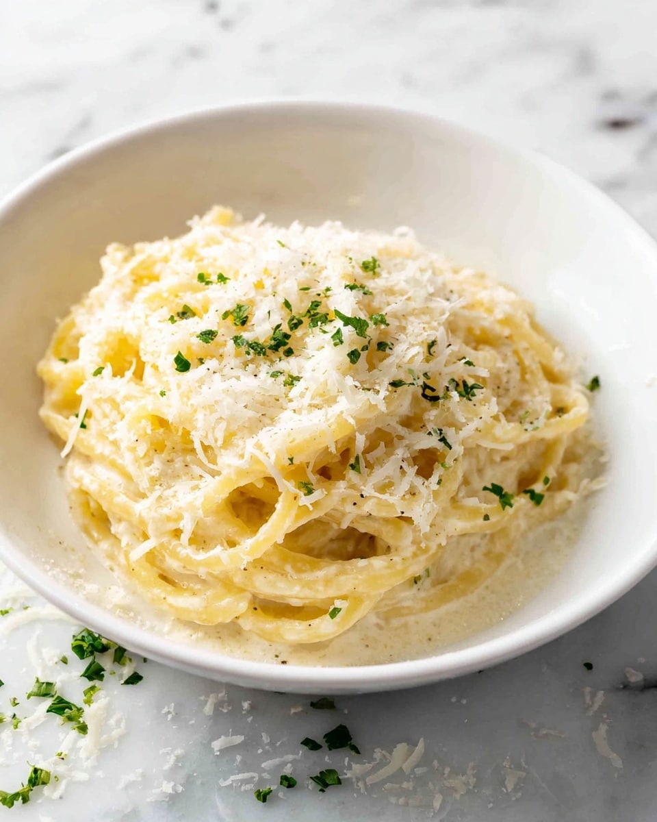 A white bowl holds a simple yet creamy pasta dish with one layer of thick, yellowish noodles coated in a smooth white sauce. The top is sprinkled with a light layer of finely grated white cheese and tiny bits of green herbs scattered across the noodles. The bowl is set on a white marbled surface with some small cheese bits and herbs around it. The overall look is fresh and inviting with soft textures and gentle coloring. photo taken with an iphone --ar 4:5 --v 7