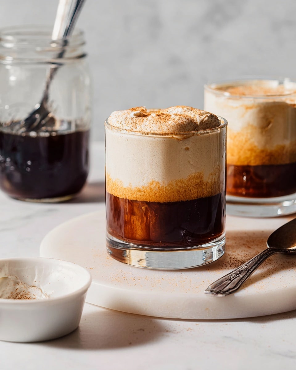 A clear glass cup shows two layers of drink: the bottom is dark brown and slightly cloudy, and the top is a thick, light cream foam with some fine brown powder sprinkled on it, sitting on a round white marble board. Behind it is another similar drink, and to the left is a large clear glass jar filled with a dark liquid and a spoon inside. On the white marbled surface in front is a small white bowl and a silver spoon with some cream residue. Soft lighting highlights the smooth and creamy textures of the drink. photo taken with an iphone --ar 4:5 --v 7