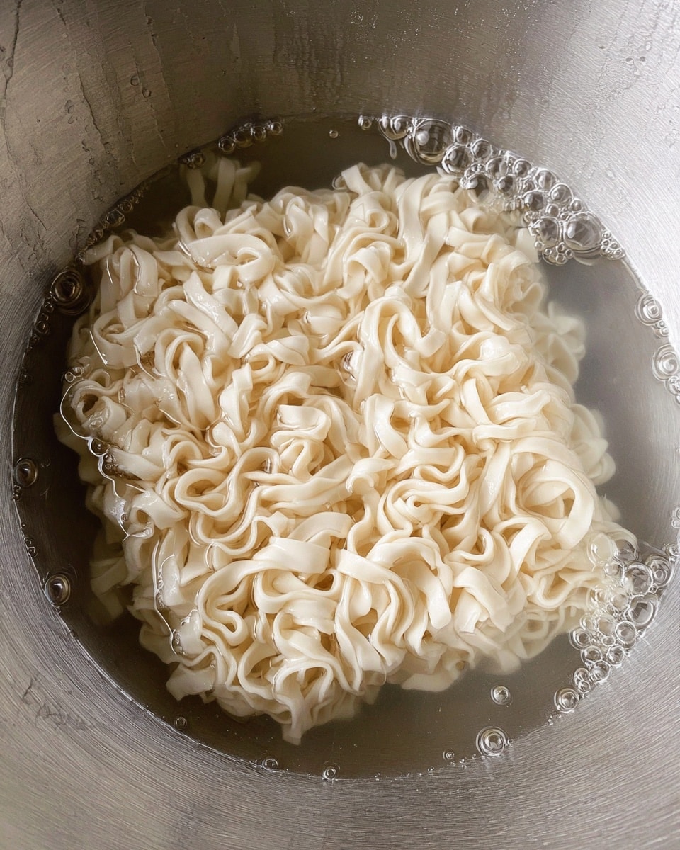 A close-up view of a single layer of uncooked instant noodle block sitting in a shiny metal pot filled with water. The noodle layer is pale cream in color with a wavy, ruffled texture that looks dense and slightly twisted. The water is clear with small bubbles clinging to the noodle surface, giving it a fresh, wet look. The metal pot has subtle reflections and marks, showing its smooth surface. The background is a white marbled texture. photo taken with an iphone --ar 4:5 --v 7