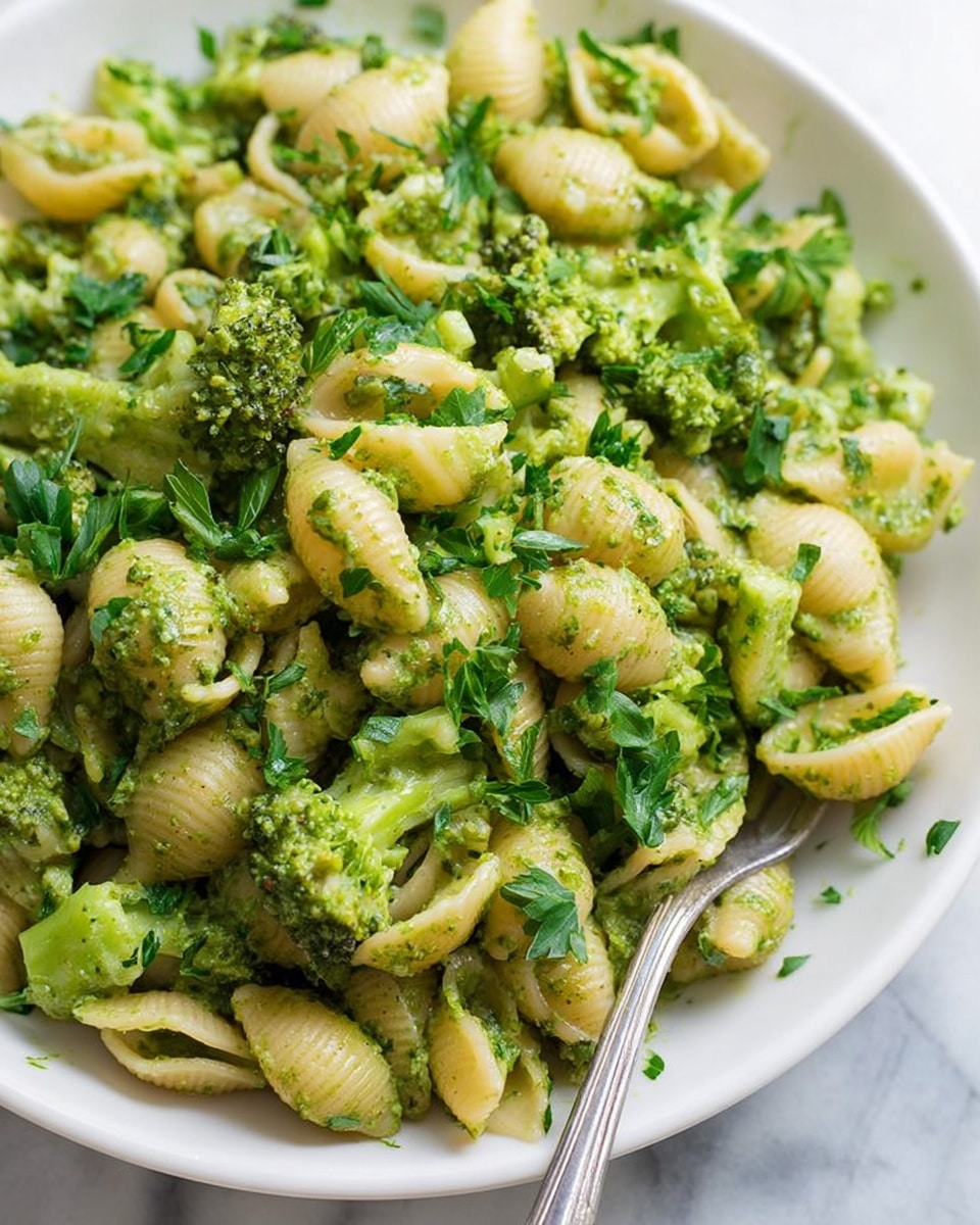 The image shows a large white plate full of small shell pasta mixed with bright green broccoli florets and chopped parsley. The pasta and broccoli are coated evenly with a green sauce that looks like pesto, giving a slightly textured and shiny appearance to the dish. The shell pasta is pale yellow while the broccoli pieces are vivid green with soft edges, creating a contrast of colors and shapes. In the lower right corner of the plate, a silver fork rests on the edge. The white marbled surface underneath makes the colors of the dish pop, and the photo is captured close up to show the fine details of the ingredients photo taken with an iphone --ar 4:5 --v 7