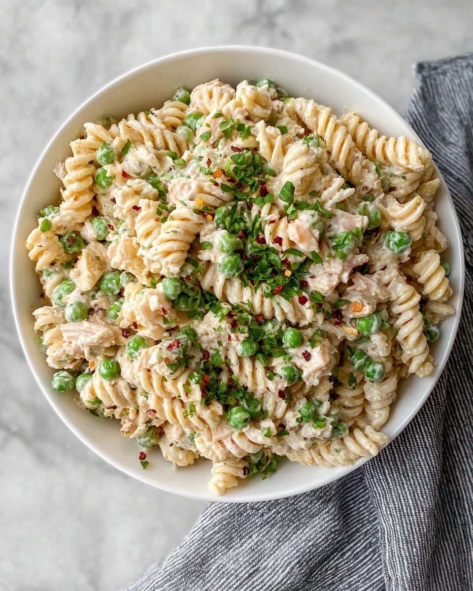 A white bowl filled with three layers of creamy pasta salad made with short, twisted rotini noodles in a light cream sauce, mixed with bright green peas and small pieces of light-colored chicken, sprinkled on top with chopped green herbs and some red pepper flakes. The creamy pasta salad has a soft, smooth texture with the peas adding small pops of green color throughout. The bowl sits on a white marbled surface with a grey striped cloth partly visible beside it. photo taken with an iphone --ar 4:5 --v 7