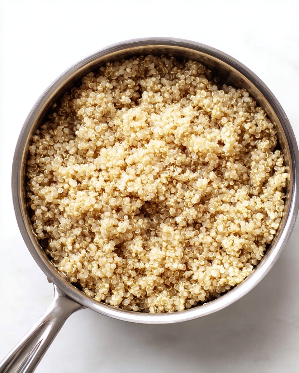 A close-up overhead view of a shiny stainless steel saucepan filled with cooked quinoa. The quinoa grains are light beige and slightly translucent with tiny white rings around each grain, looking fluffy and soft. The saucepan handle extends downward toward the bottom left corner, resting on a clean white marbled surface. The image shows only one layer of quinoa filling the pan evenly to the edges. photo taken with an iphone --ar 4:5 --v 7