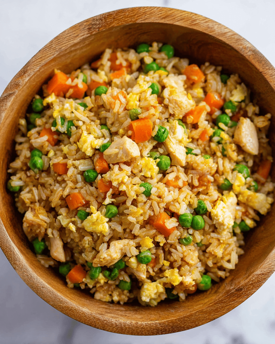 A close-up of a wooden bowl filled with fried rice showing a mix of light brown rice grains, bright green peas, small orange carrot cubes, and small yellow scrambled egg pieces evenly spread throughout. There are also chunks of light-colored cooked chicken mixed in. The bowl sits on a white marbled surface, and the texture of each ingredient is clear, with the rice grains appearing soft and slightly shiny, the peas looking smooth and round, and the carrot cubes firm. Photo taken with an iphone --ar 4:5 --v 7