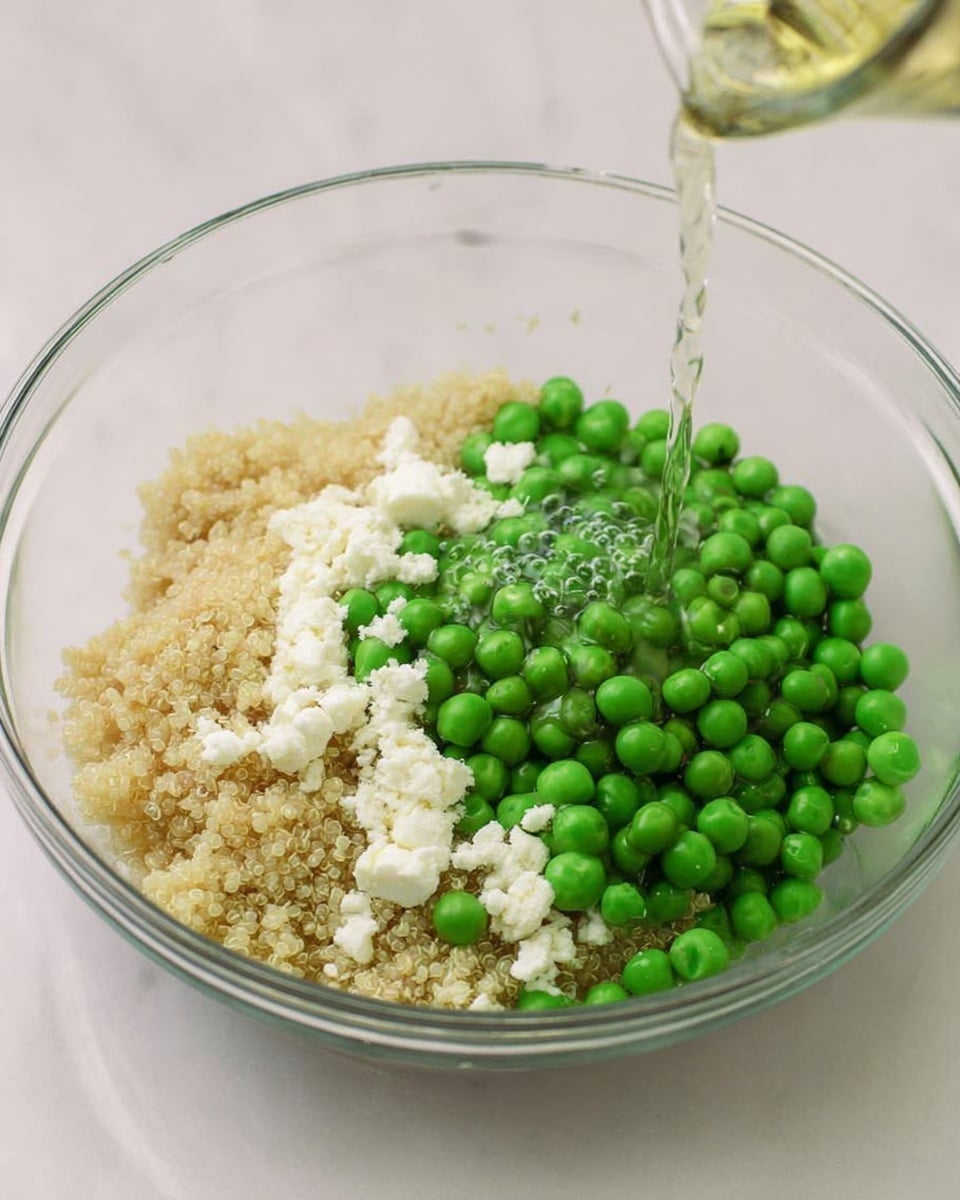In a clear glass bowl on a white marbled surface, there are three main layers: on the right, a large pile of bright green peas, smooth and round; on the left side, light beige cooked quinoa with a grainy texture; and scattered across the quinoa, crumbled white cheese pieces. A clear liquid is being poured over the ingredients from above, with a gentle stream that creates small ripples where it lands. The bowl is round and transparent, showing all the layers clearly. photo taken with an iphone --ar 4:5 --v 7