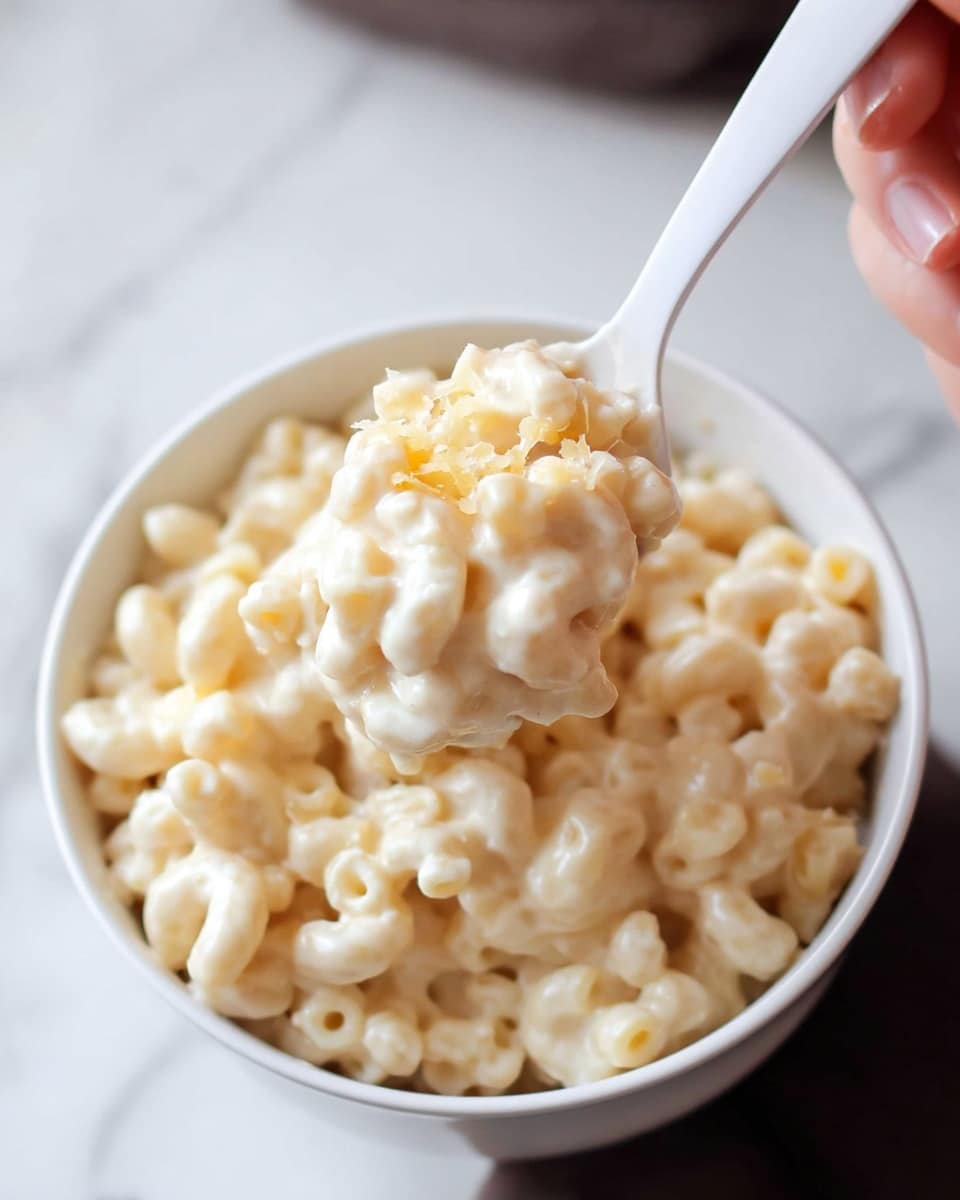 A close-up image of a white bowl filled with creamy macaroni and cheese, showing multiple small macaroni noodles fully coated in a smooth, thick white cheese sauce. The macaroni looks soft and round, with a few delicate cheese crumbs sprinkled on top. A white plastic spoon is lifting a scoop of macaroni from the bowl, held by a woman's hand that is slightly blurred in the background. The surface below the bowl is a white marbled texture, making the creamy dish stand out clearly. Photo taken with an iphone --ar 4:5 --v 7