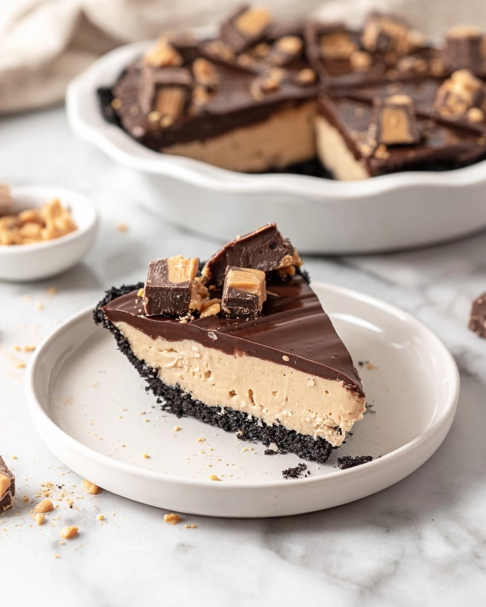 A slice of pie is placed on a white plate with a white marbled background. The pie has three layers: the bottom crust is dark and crumbly, the middle layer is a smooth light beige filling, and the top layer is a shiny dark brown chocolate glaze. On top of the pie slice, there are pieces of chocolate and peanut butter candies scattered, adding texture and color contrast. In the background, the rest of the pie in a white dish is visible, showing the same layers and candy decoration on each cut slice. There are small crumbs and bits around the plate and on the surface. Photo taken with an iphone --ar 4:5 --v 7