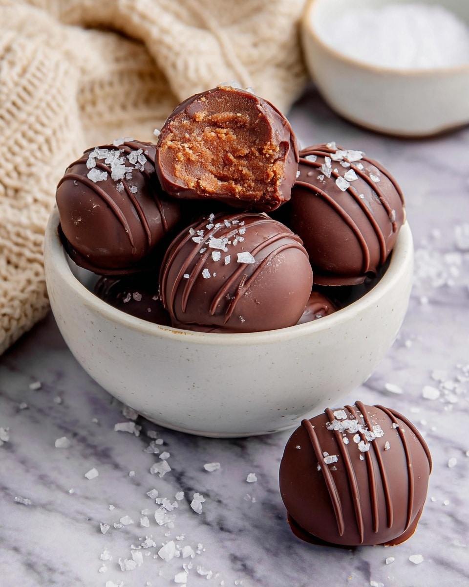 A small white bowl filled with six round chocolate truffles covered in smooth dark chocolate, each decorated with thin chocolate lines and topped with coarse flakes of sea salt. One truffle is cut open on top, showing a light brown, crumbly, and slightly moist inside layer, while another whole truffle rests outside the bowl on a white marbled surface scattered with extra salt crystals. The background shows a blurred beige knitted fabric and a white bowl with salt. Photo taken with an iphone --ar 4:5 --v 7