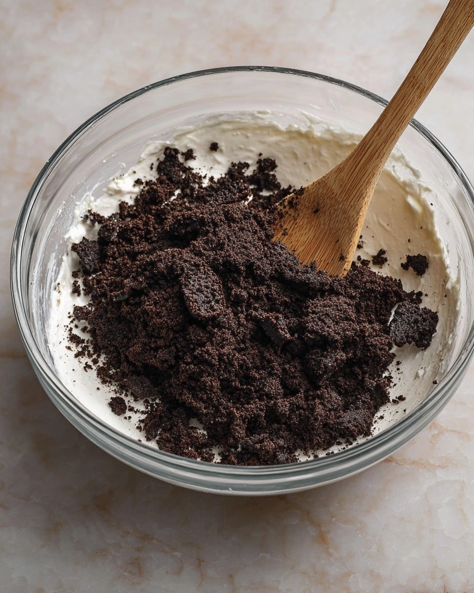 A clear glass bowl sits on a white marbled surface filled with a thick white creamy mixture as the base layer. On top of this creamy layer, there is a large pile of crushed dark brown cookies with visible chunks and fine crumbs. A wooden spoon with a smooth, light brown handle is standing upright in the bowl, partially buried in the layers. The scene captures the moment just before mixing the cookie crumbs into the creamy base. Photo taken with an iphone --ar 4:5 --v 7