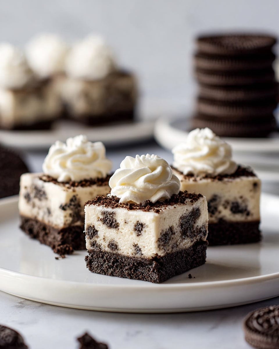 Three small square dessert pieces sit on a white plate with a white marbled background. Each piece has two layers: a thick bottom layer of dark chocolate cookie crumbs and a thicker top layer of creamy white filling mixed with dark cookie bits. On top of each dessert square is a small dollop of smooth white whipped cream with a swirl texture. In the background, stacks of dark chocolate cookies and another white plate with similar desserts are slightly out of focus. Photo taken with an iphone --ar 4:5 --v 7