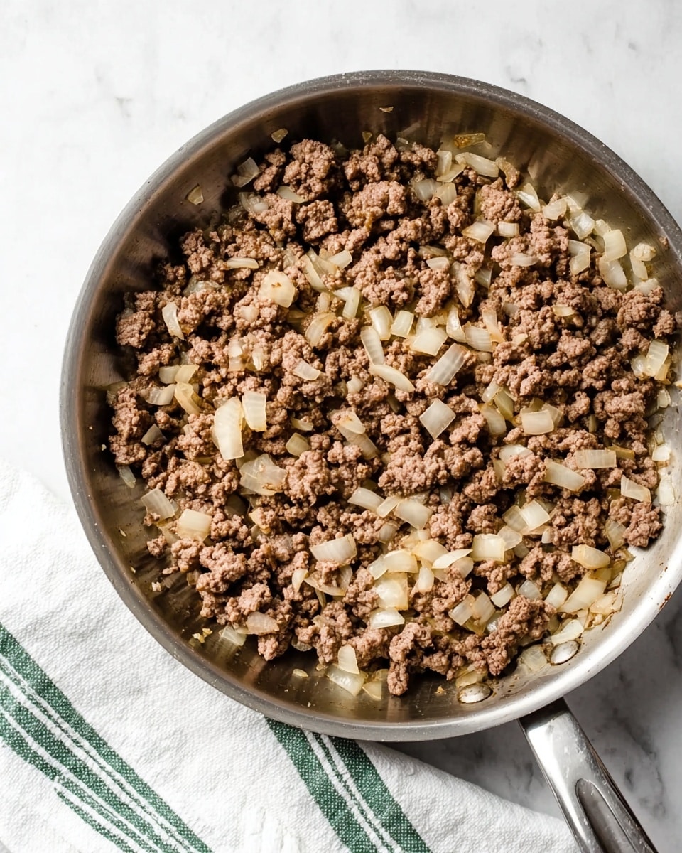 A stainless steel pan filled with cooked ground beef mixed with soft, translucent chopped onions spread evenly across the pan. The beef is crumbly with shades of brown and a little pink, and the onions are small white chunks with a slight shine. The pan handle is visible on the right side and there is a white cloth with green stripes placed near the top-right edge of the pan. The surface under the pan has a white marbled texture photo taken with an iphone --ar 4:5 --v 7