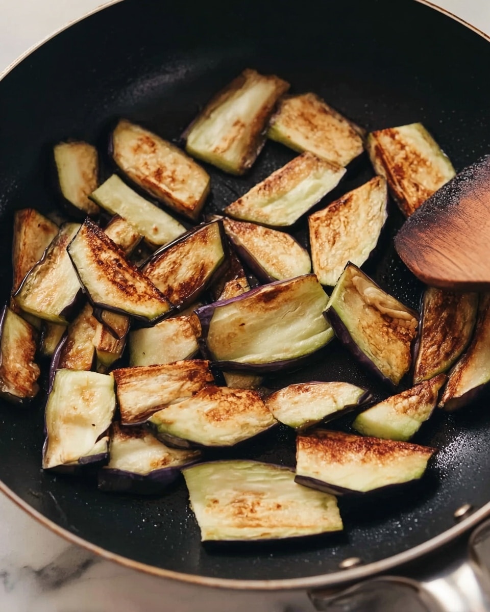 A frying pan holds many pieces of cooked eggplant slices. Each slice has a browned, slightly crispy-looking surface with a soft pale green and creamy white inside. The eggplants are uneven in shape, mostly wedge-like, scattered evenly across the pan's dark surface. The pan is on a white marbled surface, and a woman's hand with a wooden spatula stirs the eggplants gently. The light shines softly, highlighting the texture and light char on the eggplant pieces photo taken with an iphone --ar 4:5 --v 7