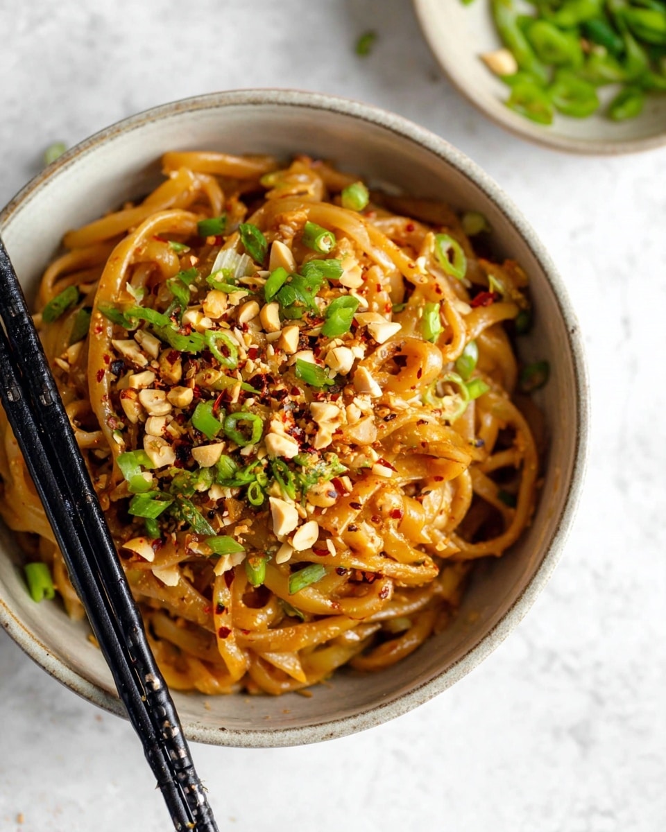 A round bowl filled with thick, light brown noodles coated in a smooth sauce, topped with chopped green onions and crushed peanuts scattered evenly over the surface; red chili flakes are sprinkled on top adding small red dots; black chopsticks rest on the left side of the bowl; the background is a white marbled texture with a small bowl of green onions blurred in the top right corner photo taken with an iphone --ar 4:5 --v 7