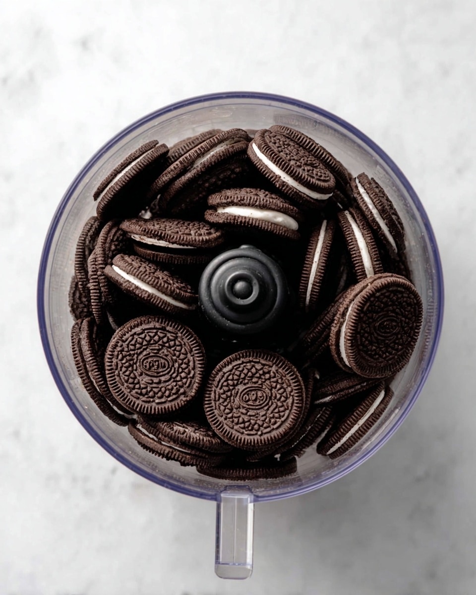 A clear food processor bowl filled with a single layer of whole chocolate sandwich cookies with white cream filling, stacked closely together. The bowl sits on a white marbled surface, and the view is directly from above, showing the detailed embossed pattern on each cookie. The central black blade mechanism is visible, surrounded by the dark brown cookies with a slightly textured surface and cream centers peeking between some of them. photo taken with an iphone --ar 4:5 --v 7