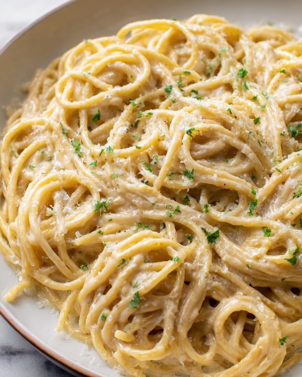 A close-up view of a large white plate filled with creamy spaghetti coated in a thick, light beige sauce. The noodles are twisted into multiple loose bundles across the plate, showing their long, smooth texture. Small green herb pieces are sprinkled evenly on top, adding a touch of color. The sauce has a slightly grainy texture, giving a rich and creamy look. The white marbled surface under the plate adds a clean and simple background. photo taken with an iphone --ar 4:5 --v 7