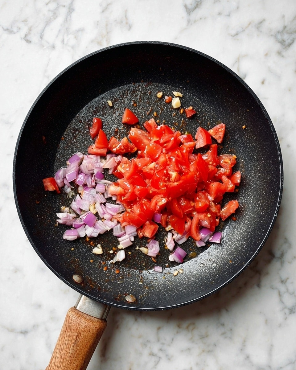 A black frying pan with a wooden handle sits on a white marbled surface, containing chopped red tomatoes in the center with small pieces of light purple onions scattered mainly to the left and bottom sides, along with a few chopped white garlic pieces near the bottom. The pan shows a light sheen of oil, giving a slightly shiny texture to the ingredients. photo taken with an iphone --ar 4:5 --v 7