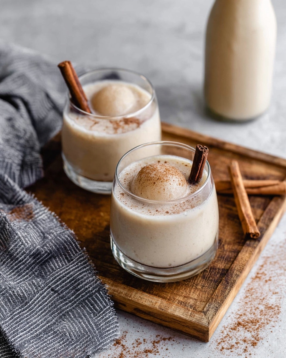 Two clear glasses sit on a wooden board with a white marbled texture beneath. Each glass holds a creamy light beige drink with a large round ice sphere inside. On top of the drink in each glass lies a dark brown cinnamon stick, and a dusting of cinnamon powder is sprinkled over the surface. Three additional cinnamon sticks rest on the wooden board beside the glasses. A gray and white striped cloth is casually placed near the board. In the blurry background, a bottle with more of the creamy drink is visible. photo taken with an iphone --ar 4:5 --v 7