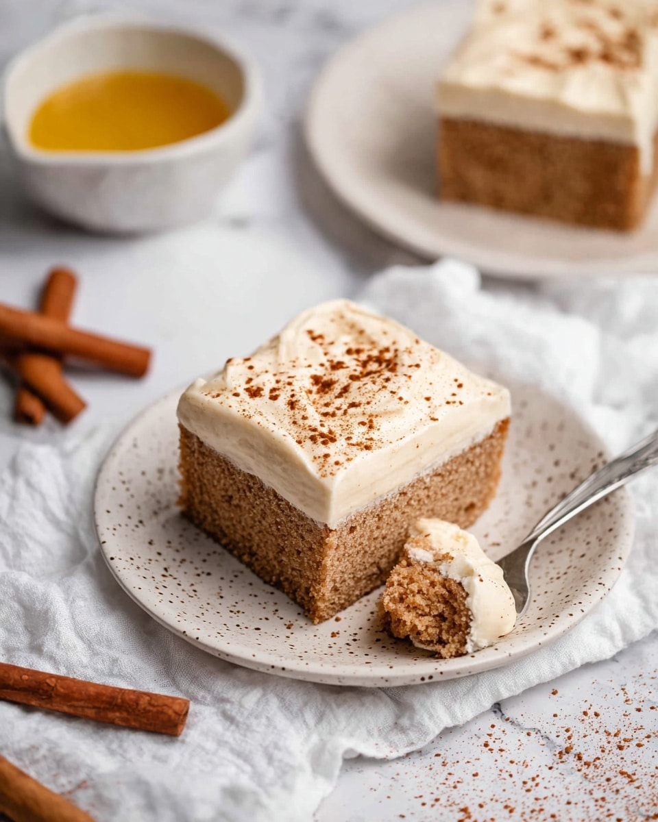 A square piece of cake with one layer of light brown spongy texture at the bottom and a smooth layer of off-white frosting on top, sprinkled lightly with brown powder. The cake is placed on a white speckled plate with a silver spoon holding a bite-sized piece of the cake with frosting, resting beside it. In the background, there is another similar square piece of cake on a white speckled plate, a small white bowl with a yellow sauce, and two cinnamon sticks on a crinkled white cloth over a white marbled surface. photo taken with an iphone --ar 4:5 --v 7