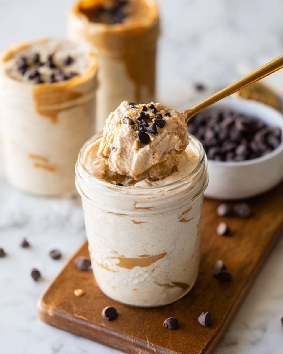 The image shows a clear glass jar filled with a creamy light beige mixture, with a swirl of golden brown peanut butter visible along the inside edges. On top of the mixture is a scoop of thick, beige creamy texture sprinkled with small dark chocolate chips. A gold spoon is scooping some of this creamy mix from the jar. The jar is placed on a wooden board with a few scattered dark chocolate chips nearby. In the background, two more jars filled with a similar creamy mixture are visible along with a small white bowl filled with dark chocolate chips. The surface underneath everything is white with a marbled texture. Photo taken with an iphone --ar 4:5 --v 7