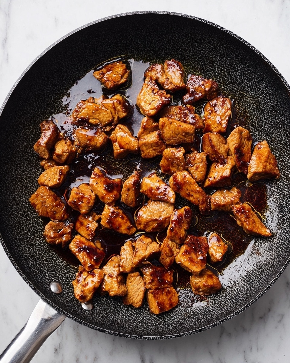 A black frying pan on a white marble surface filled with small, cooked pieces of meat that are browned with a shiny, dark sauce coating them unevenly. The meat pieces vary in size and shape, some with a light golden color while others have darker charred spots. The pan has a textured hexagonal pattern inside and a metal handle extending to the left. There is some oil pooled around the meat pieces, reflecting light. photo taken with an iphone --ar 4:5 --v 7