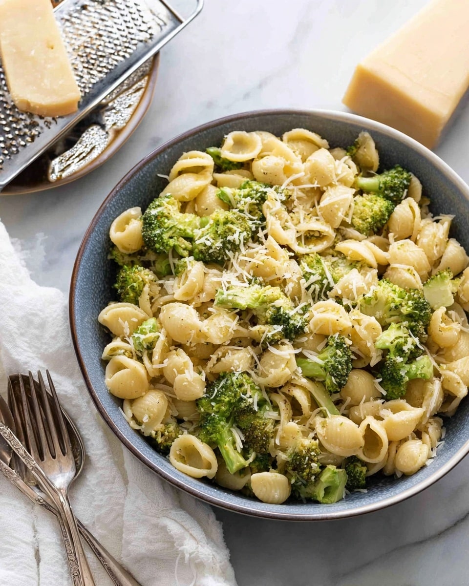 A large bowl filled with small shell pasta mixed with bright green broccoli florets, all topped with a light sprinkling of finely grated pale yellow cheese. The pasta and broccoli have a soft, slightly shiny texture, with visible small herbs or seasoning mixed in. The bowl is round with a blue-gray outside and white inside, placed on a white marbled surface, next to a white cloth and two forks. Nearby, a block of cheese rests on a metal grater. Photo taken with an iphone --ar 4:5 --v 7