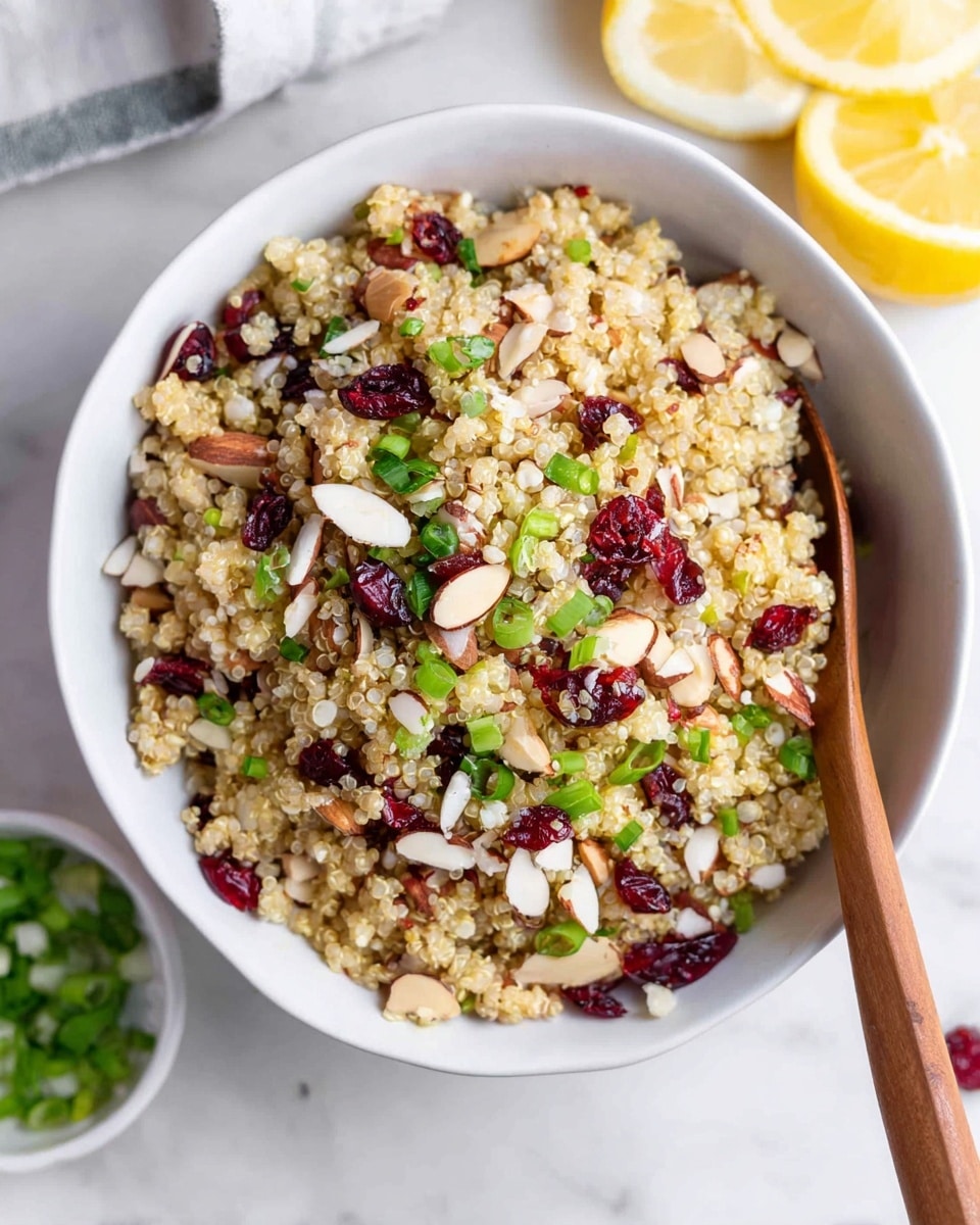 The image shows a white bowl filled with a mixed salad that has three main layers visible: a light tan base of cooked quinoa with a fluffy texture, scattered bright red dried cranberries, and almond slices on top adding light brown and off-white tones with a crunchy texture. Green chopped scallions are spread over the dish, adding fresh green color and texture. There is a wooden spoon resting inside the bowl on the right side. The bowl sits on a white marbled surface, and around it there are lemon halves and a small white bowl with more scallions. The photo taken with an iphone --ar 4:5 --v 7