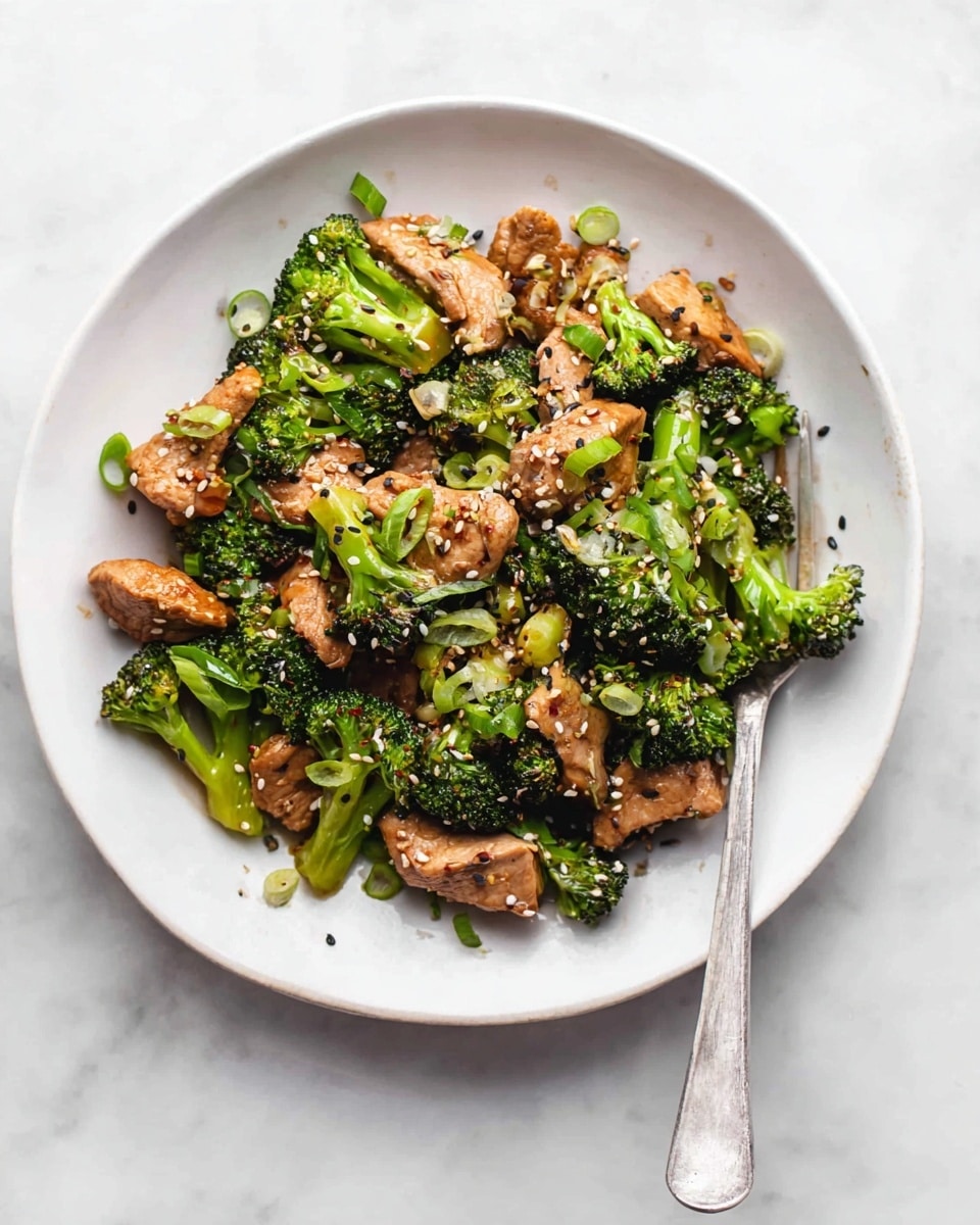A white round plate sits on a white marbled surface, filled with a stir-fry dish that has two main layers. The bottom layer contains bright green broccoli florets with a slightly rough texture and small stems. On top and mixed through are medium-sized pieces of cooked meat in light brown color, with a slightly shiny and soft texture. The dish is garnished with thin slices of green onion scattered over, adding a fresh green touch, and sprinkled white and black sesame seeds. A silver fork is placed on the right side of the plate, partially resting on the food. Photo taken with an iphone --ar 4:5 --v 7