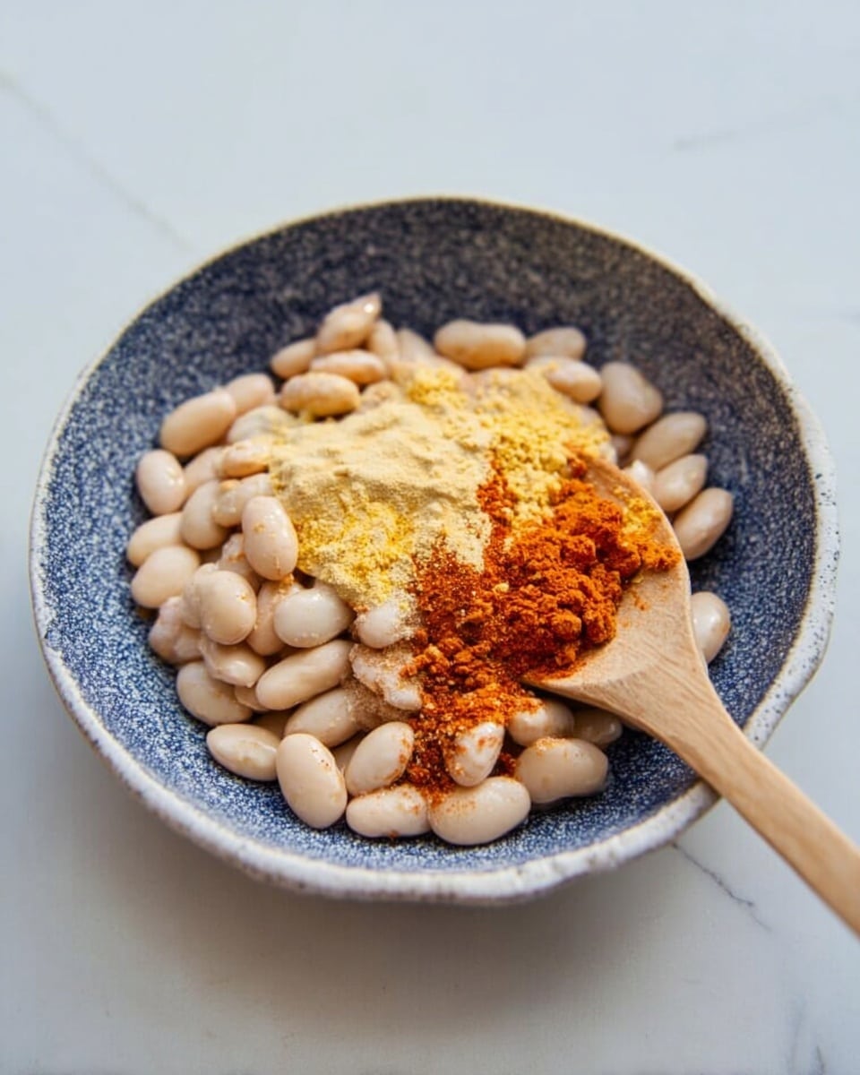 A shallow white bowl with a blue speckled inside holds a layer of white beans spaced evenly at the bottom. On top of the beans, there is a mix of light yellow powder and reddish-brown powder sprinkled unevenly. A wooden spoon sits inside the bowl on the right side, partially covered by the beans and powders. The bowl rests on a white marbled surface. photo taken with an iphone --ar 4:5 --v 7