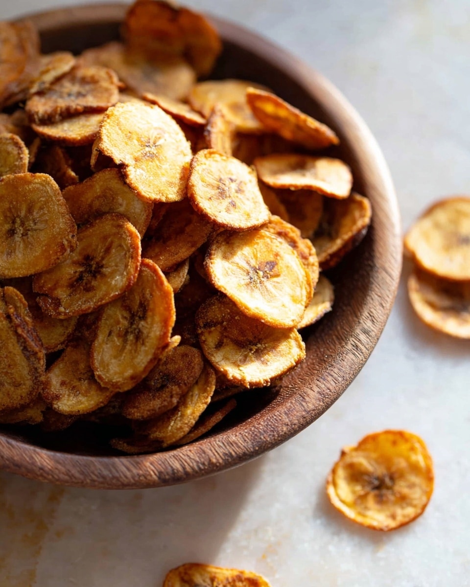 A close-up image of round, thin banana chips piled high inside a rustic wooden bowl. The chips are golden brown with varying shades of caramel and light tan, showing a crispy texture with some darker caramelized spots. A few chips are scattered around the bowl on a white marbled surface, adding to the casual and natural feel. The lighting highlights the crunchy texture and the rich colors of the banana slices. photo taken with an iphone --ar 4:5 --v 7