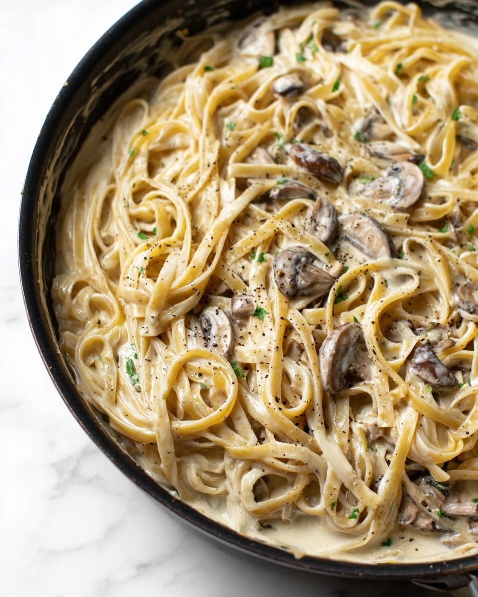 A black pan filled with creamy mushroom pasta sits on a white marbled surface. The dish has one main layer of light golden fettuccine noodles coated in a thick, pale cream sauce with visible small green herb bits. Scattered throughout are thin slices of brown mushrooms, some partially covered by the sauce. The noodles are gently twisted over each other, with a slight shine on the creamy surface, and tiny black pepper specks are sprinkled on top. Photo taken with an iphone --ar 4:5 --v 7