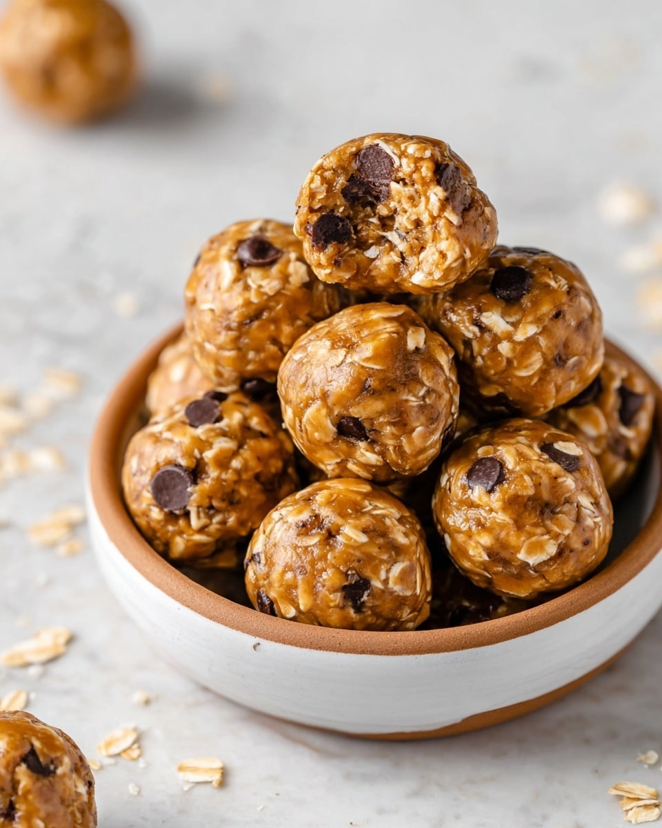 A bowl full of round, small energy bites stacked on top of each other, each bite showing a textured mix of light brown, creamy peanut butter with visible oats and dark chocolate chips mixed inside, one bite at the top has a small bite taken out of it, revealing a soft, dense inside of oats and chocolate. The bowl is white with a brown rim and sits on a white marbled surface with a few oats scattered around. Photo taken with an iphone --ar 4:5 --v 7
