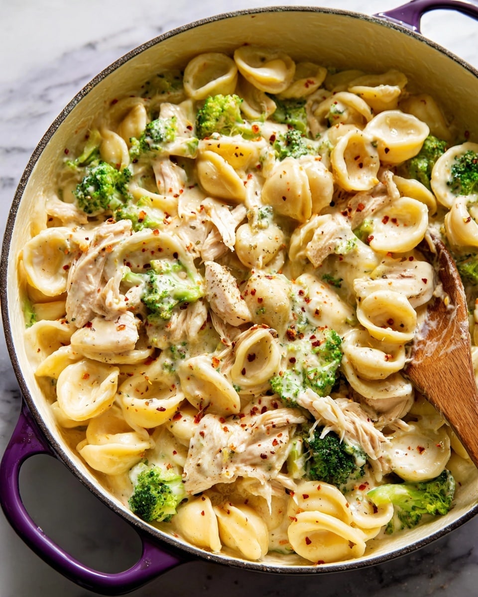 A close-up view of a creamy pasta dish in a white pot with purple handles, featuring three main layers: light yellow orecchiette pasta shells thickly coated in a glossy, creamy white sauce with hints of green herbs; shredded pieces of light brown chicken mixed throughout; and small bright green broccoli florets evenly spread, adding a pop of color. The dish is sprinkled lightly with black pepper and red pepper flakes, and a wooden spoon is resting on the side, partially covered by the pasta. The pot is set on a white marbled surface. Photo taken with an iphone --ar 4:5 --v 7