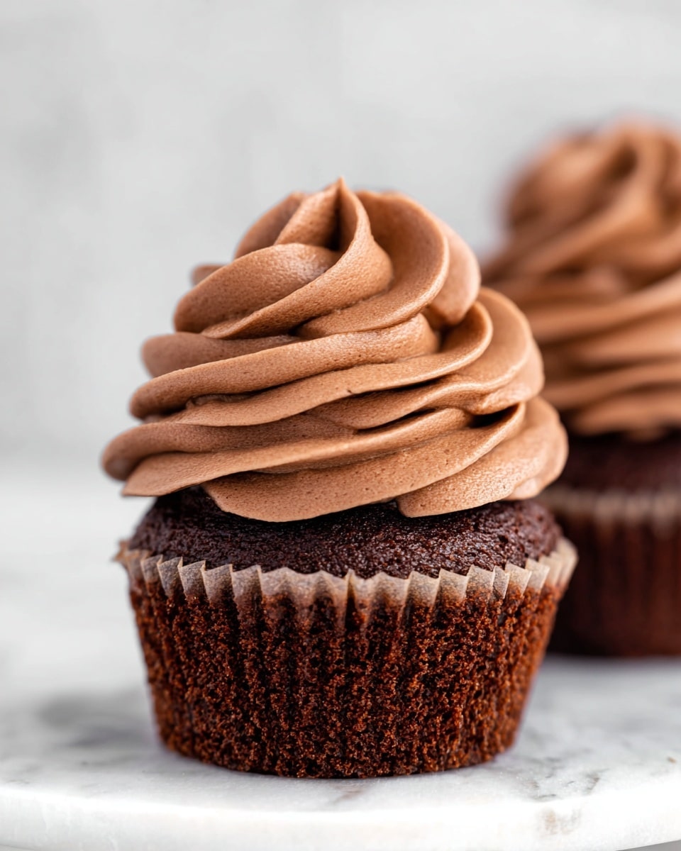 The image shows a close-up of a chocolate cupcake with one visible layer of dark brown cake topped with a thick swirl of smooth, light brown chocolate frosting. The frosting is piped in wide, curved ribbons that stack tall and look creamy with a soft texture. The cupcake sits in a slightly crinkled brown paper liner and is placed on a white marbled surface. Another similarly decorated cupcake is partially visible blurred in the background. photo taken with an iphone --ar 4:5 --v 7