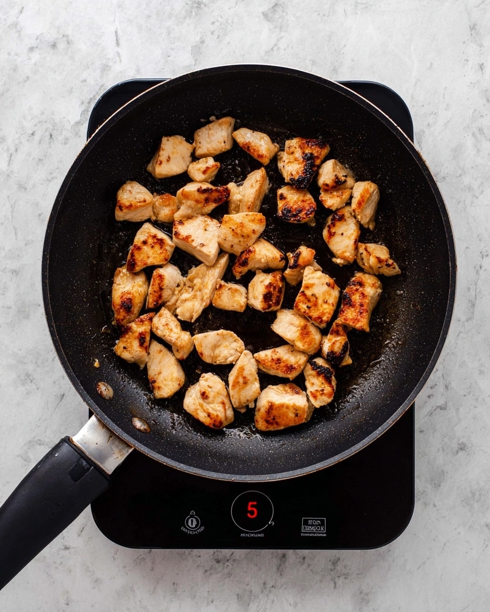 A black frying pan filled with about twenty small pieces of browned cooked chicken with different shades of golden brown and slight charring. The pan is resting on a black electric hot plate with a dial knob showing number 5. The background and surface beneath the hot plate have a white marbled texture. The cooked chicken pieces vary in size and shape and are evenly spaced around the pan. Photo taken with an iphone --ar 4:5 --v 7