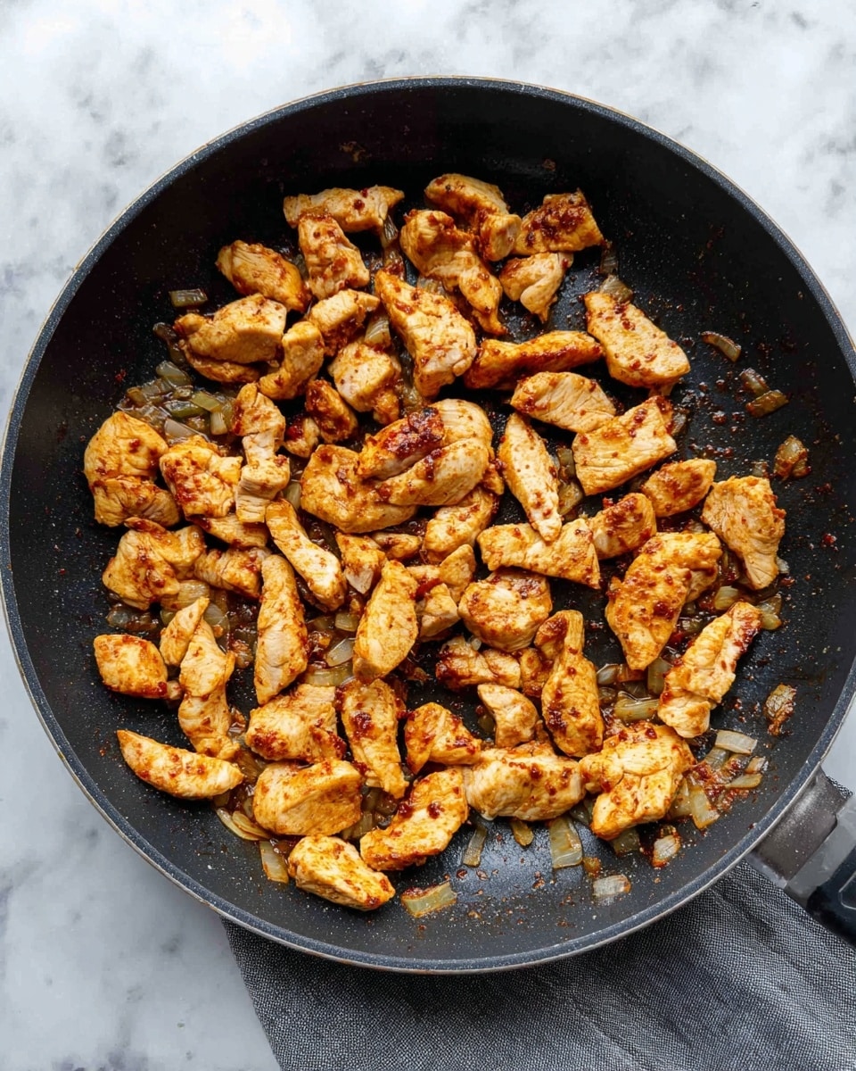 In a black frying pan, there are many pieces of cooked chicken with a golden-brown color, mixed with small pieces of cooked onions that look slightly translucent and soft. The chicken is spread unevenly, showing some darker seared spots. The pan is placed on a white marbled surface and a corner of a grey cloth is visible below the pan. The chicken pieces are small and irregular in shape, covering most of the pan's surface. Photo taken with an iphone --ar 4:5 --v 7