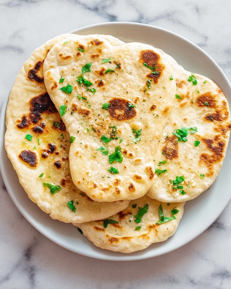 Three pieces of flatbread stacked on a white plate, each flatbread showing uneven golden-brown spots from cooking, with a soft, slightly fluffy texture; scattered on top are small green leaves adding bright color contrast. The flatbreads vary slightly in shape, with the top one more irregular, and all have a light dusting of seasoning visible. The plate sits on a white marbled surface. photo taken with an iphone --ar 4:5 --v 7