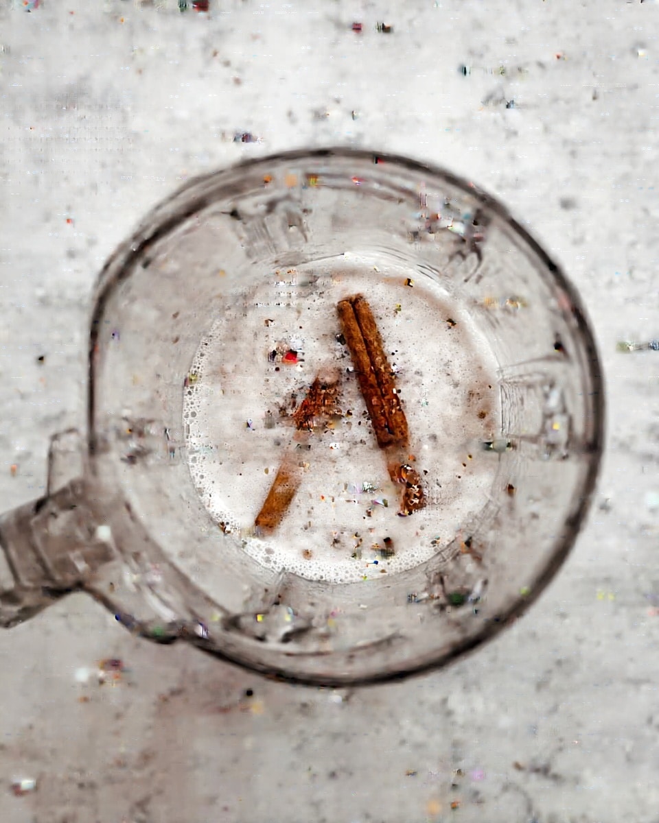 The image shows a clear blender container filled with white liquid, with two brown cinnamon sticks floating on the surface inside. The blender is placed on a white marbled surface, and the liquid has small bubbles around the cinnamon sticks. The shot is taken from above, focusing on the contents inside the blender. photo taken with an iphone --ar 4:5 --v 7