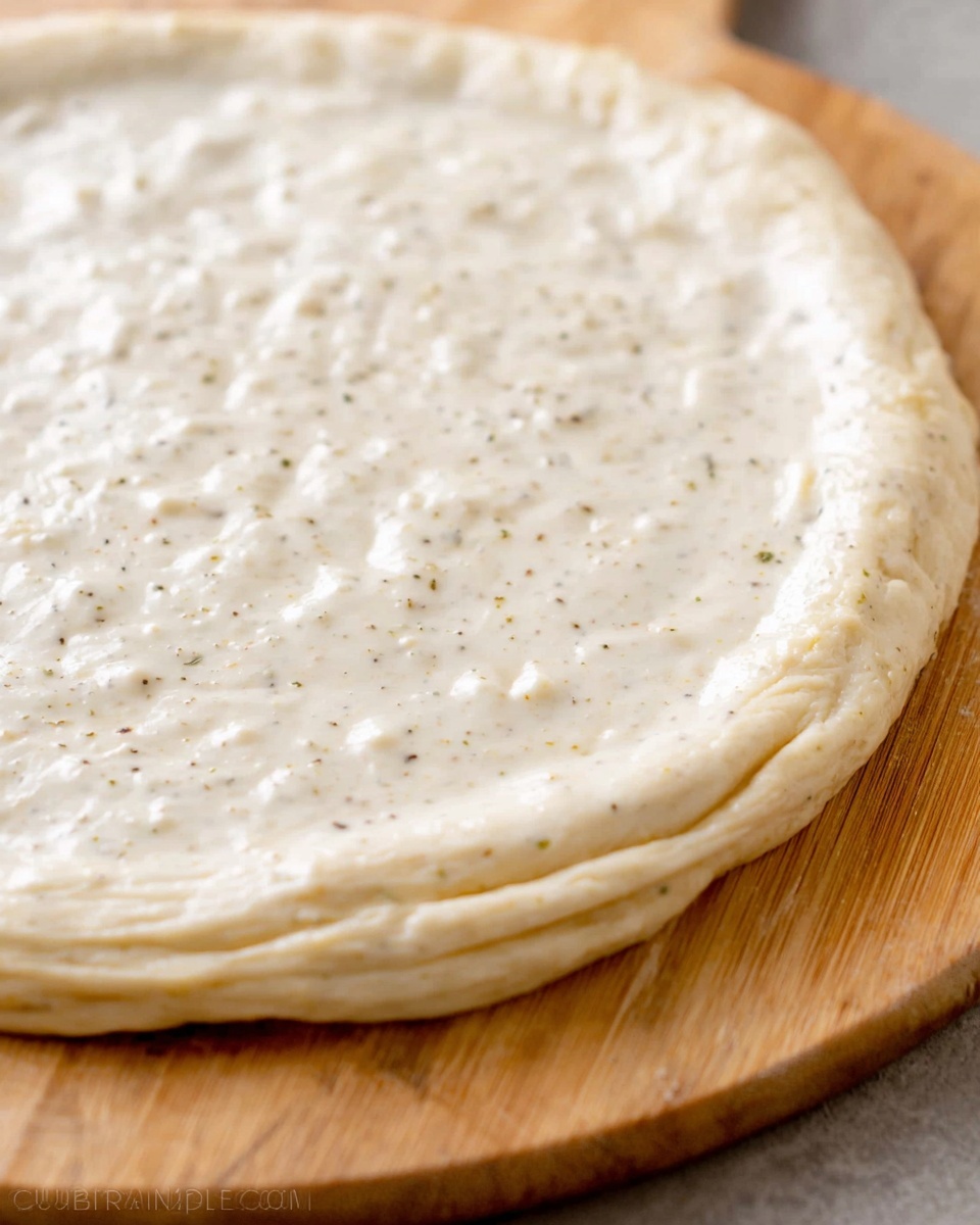 A close-up view of a round pizza dough laid flat on a wooden board with a creamy white sauce evenly spread across the surface, showing a slightly thick texture with small bubbles and specks of seasoning. The dough appears soft and pale, with faint ridges visible around the edges, indicating the rustic handmade preparation. The wooden board underneath has a warm, natural grain that contrasts with the smooth sauce topping. photo taken with an iphone --ar 4:5 --v 7